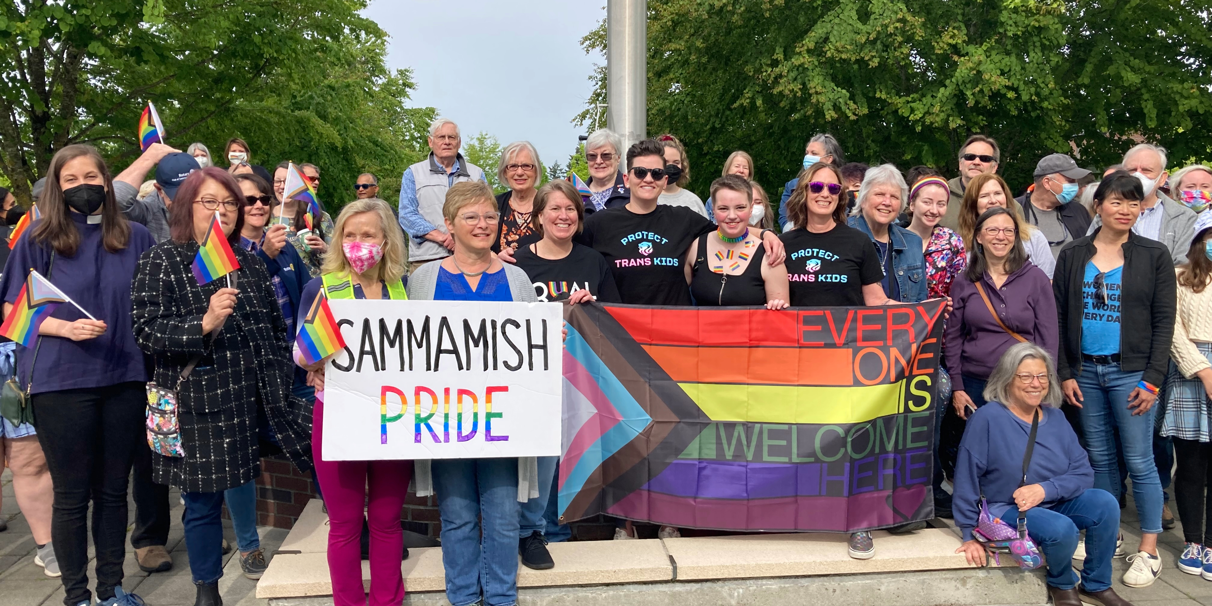 A large group of people gather outside, smiling and holding rainbow flags. Several hold signs, including one reading - SAMMAMISH PRIDE - � and another rainbow banner that says - EVERYONE IS WELCOME HERE. - �.