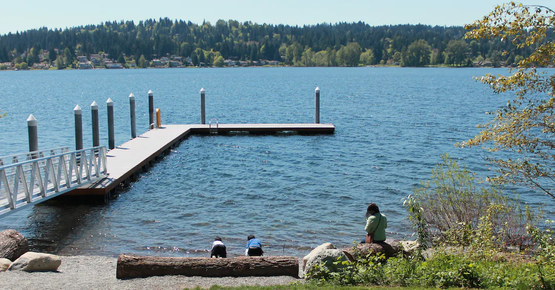 Three people sit on the shore of Lake Sammamish at Sammamish Landing Park near the dock.