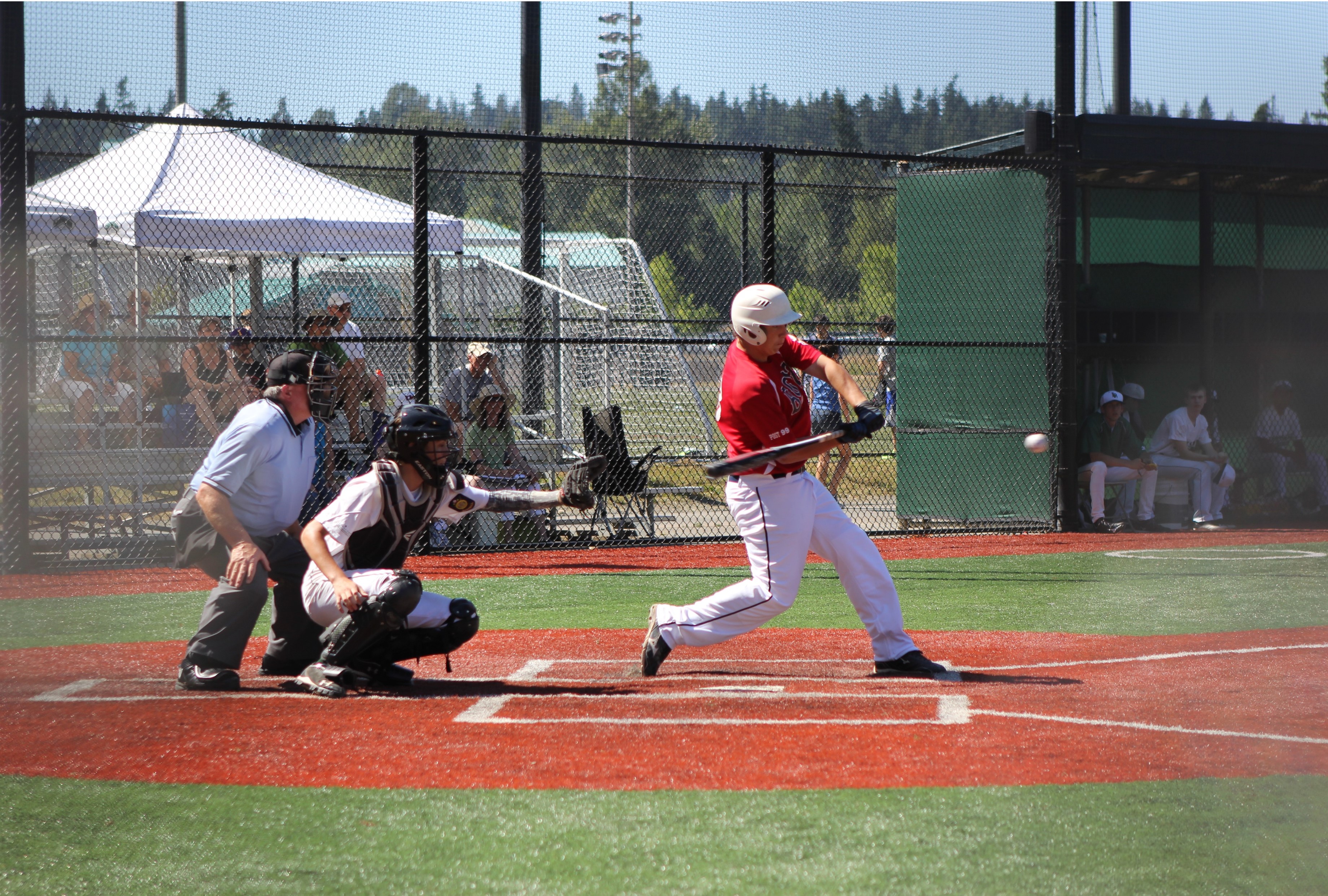 Baseball player in red shirt and white pants takes a swing with bat at Eastlake Community Sports Field 3 in Sammamish with catcher and umpire behind him.