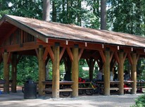 A small group gathers under the roof of a wooden picnic shelter with several picnic tables. A garbage can and barbeque grill stand next to it and it is surrounded by trees.